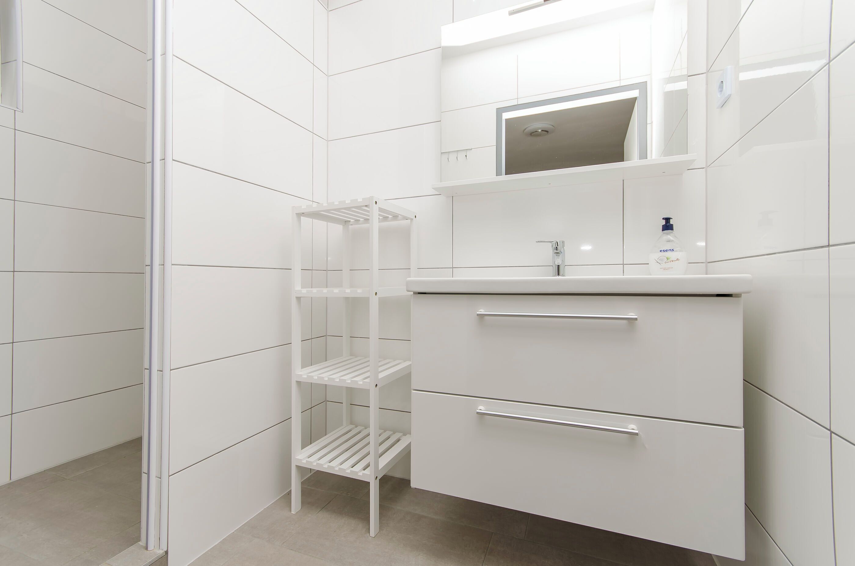 Casa de banho moderna com azulejos brancos, toucador branco com espelho e zona de duche com unidade de prateleiras brancas.