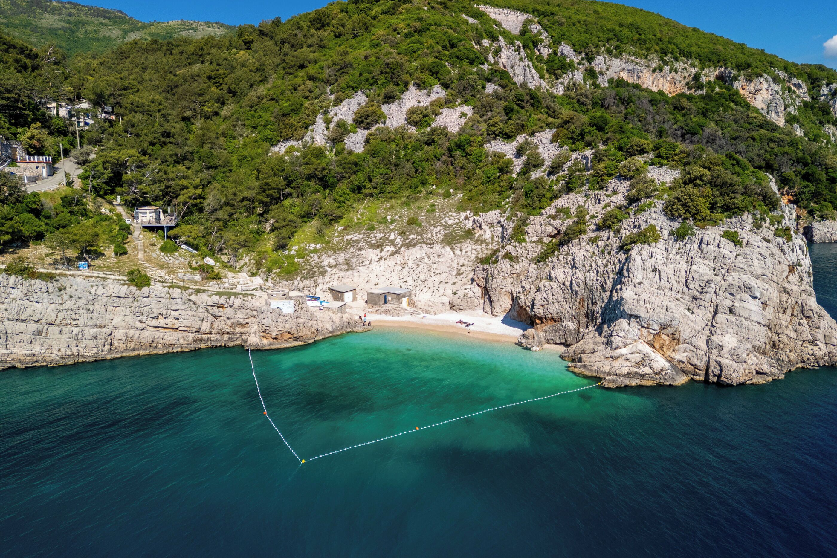 A coastal area with a rocky beach, clear turquoise waters, and a small building near the shore.