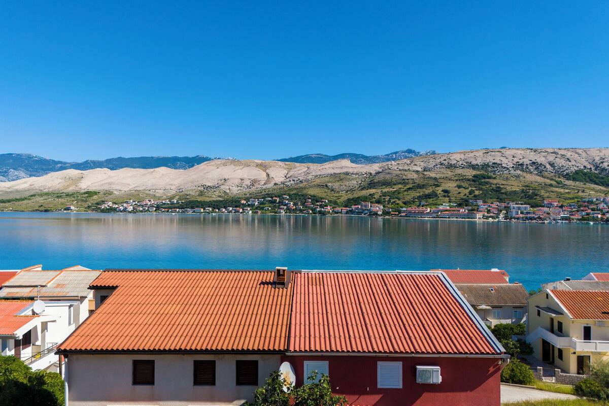 A coastal town with red-roofed buildings, a clear blue lake, and mountains in the background.