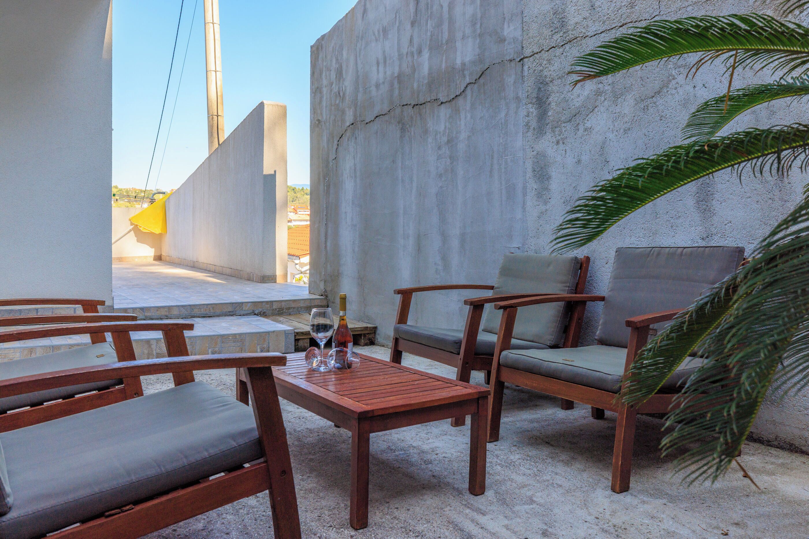 A patio with wooden furniture, a table with glasses and a bottle, and a palm tree.
