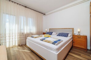 A neatly made bed with a wooden headboard, a bedside table with a lamp, and a window with sheer curtains. - Apartment Anita (Crikvenica)