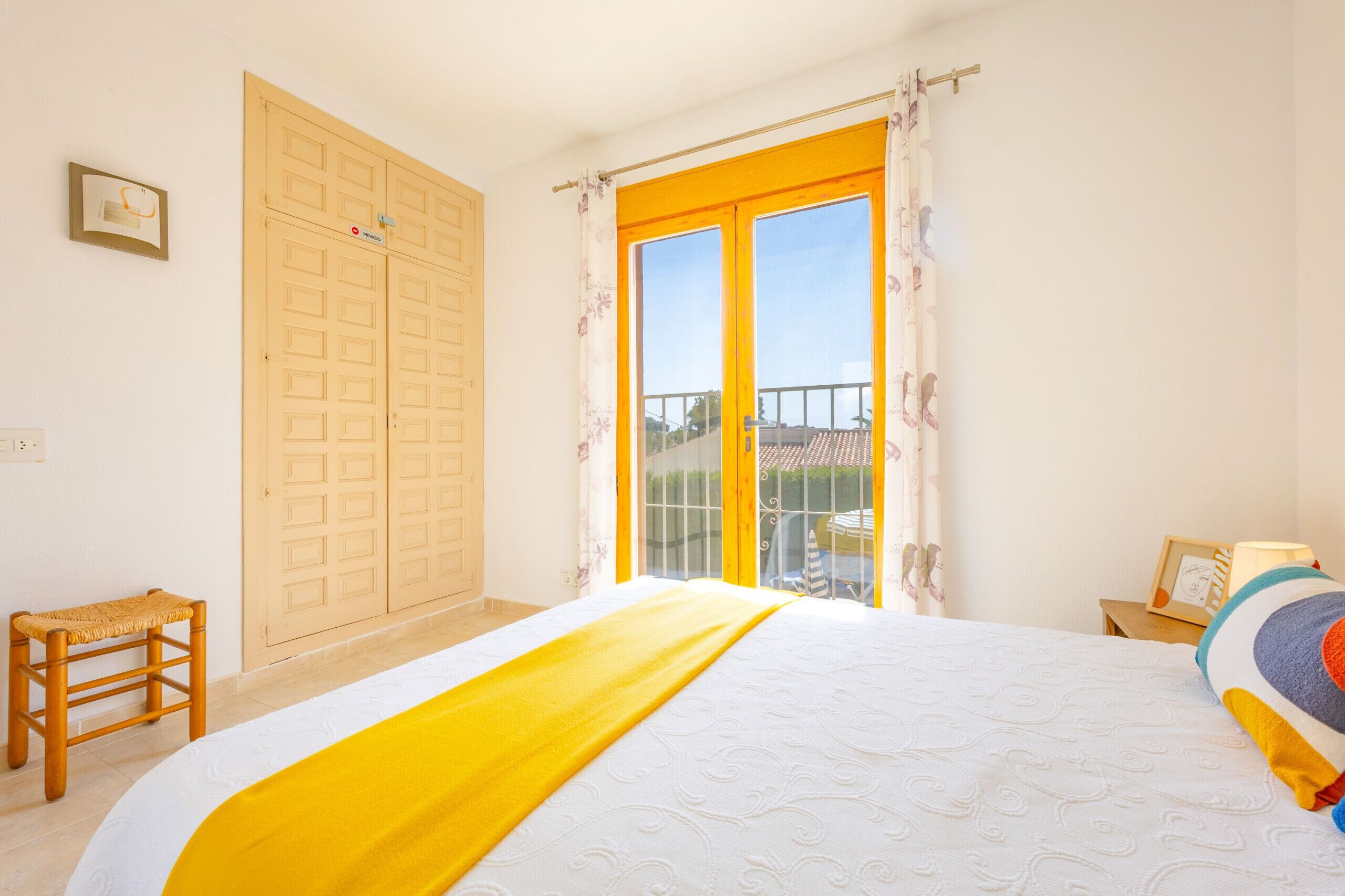 A bedroom with a bed, a wooden stool, a wardrobe, and a balcony with a view.
