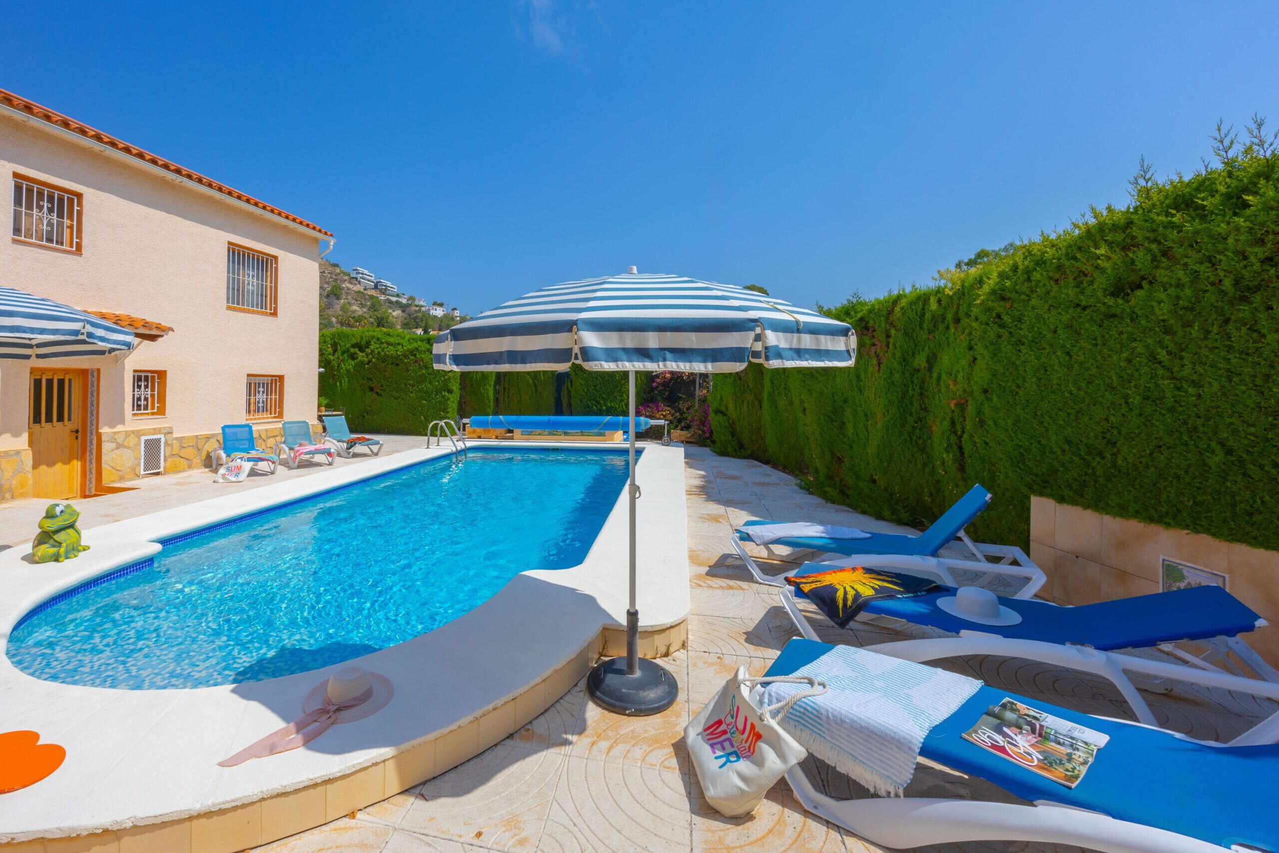 Piscina com espreguiçadeiras e guarda-sóis, cercada por um conjunto de arbustos bem cuidados e um edifício com telhado em azulejos.