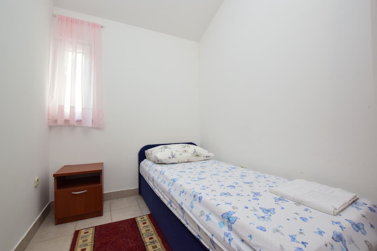 A small, single-bed room with a wooden nightstand, a red rug, and a window with a pink curtain.