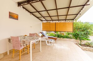 Un'area salotto esterna riparata, con tavolo e sedie, una panca imbottita e vista su piante e un edificio sullo sfondo.