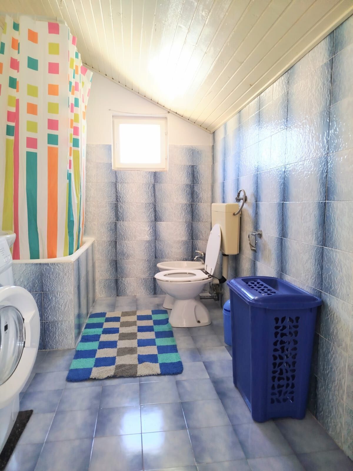 A bathroom with a checkered floor mat, a toilet, a washing machine, and a blue laundry basket.