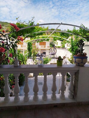 A balcony with a white balustrade, potted plants, a hanging chair, and a view of a building with a balcony. - Apartments Rose (Sibenik)
