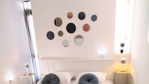 Une chambre à coucher bien rangée, avec un lit, des oreillers, un miroir fixé au mur et une petite table de chevet avec une lampe.