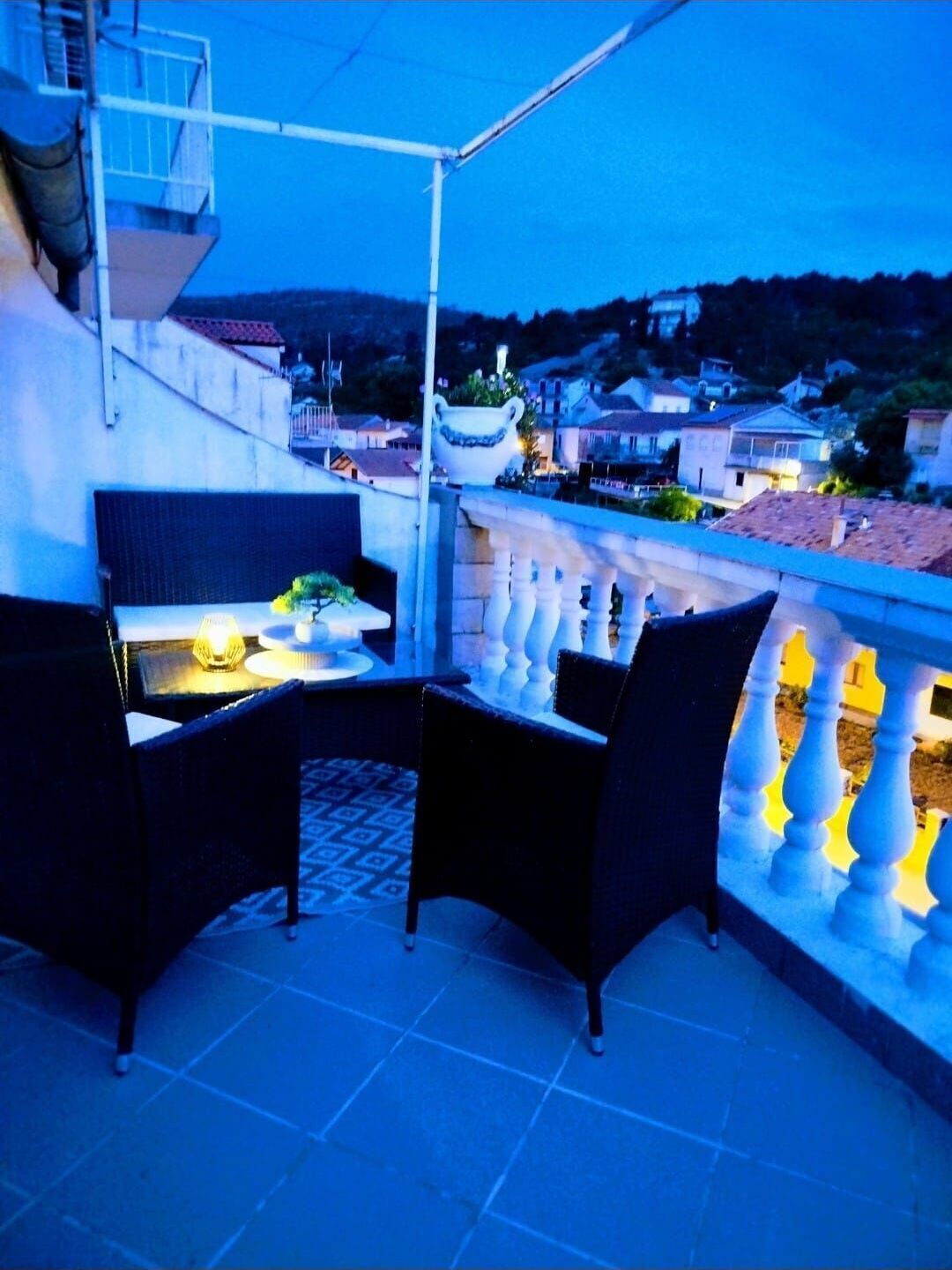 A balcony with two chairs and a table, overlooking a residential area with houses and a hill in the background.