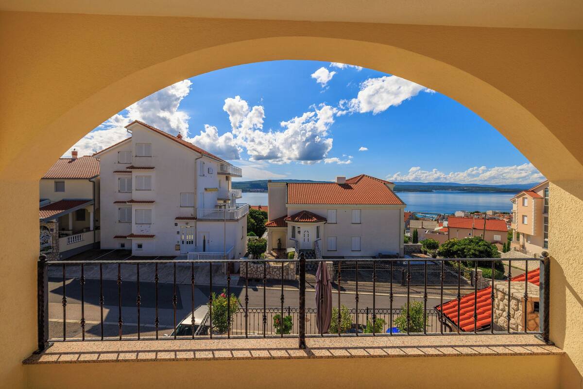 Ein Balkon mit Blick auf eine Küstenstadt, mit Häusern mit roten Ziegeldächern und blauem Himmel.