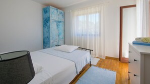 A bedroom with two beds, a wardrobe, a desk, and a window with sheer curtains.