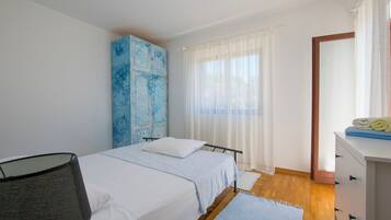 A bedroom with two beds, a wardrobe, a desk, and a window with sheer curtains.