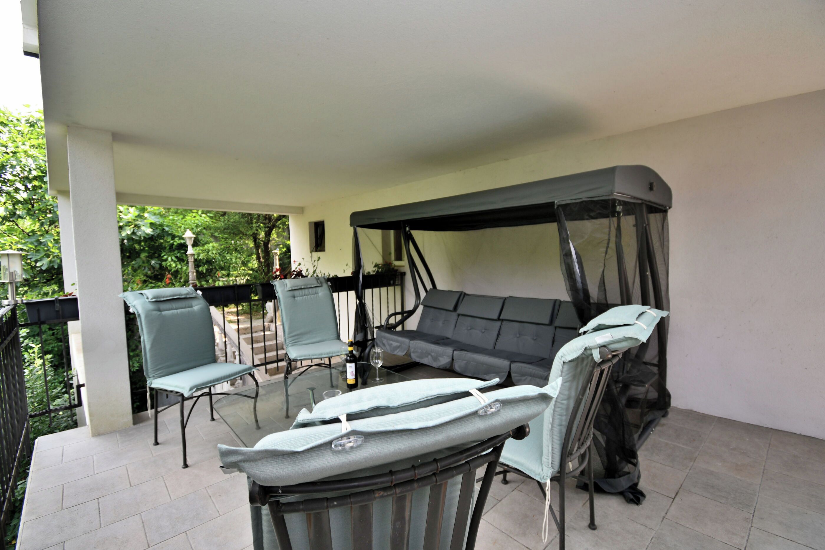 A covered outdoor seating area with a sofa and chairs, a table with a bottle and glasses, and a canopy.
