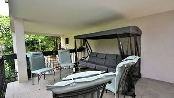 A covered outdoor seating area with a sofa and chairs, a table with a bottle and glasses, and a canopy.