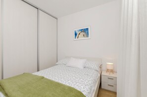 A bedroom with a bed, a nightstand with a lamp, a green blanket, a white pillow, and a framed picture on the wall. - Apartment Rose Garden (Rovinj)