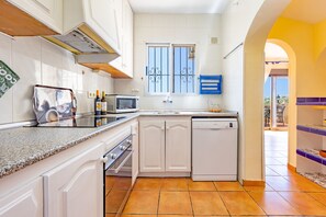A compact kitchen with white cabinets, a microwave, a dishwasher, and a window with decorative bars. - Villa Ecalinda - Plusholidays (Calpe)
