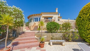 Una villa de estilo mediterráneo con una escalera de piedra que lleva a un balcón, rodeada de plantas en macetas y una tumbona.