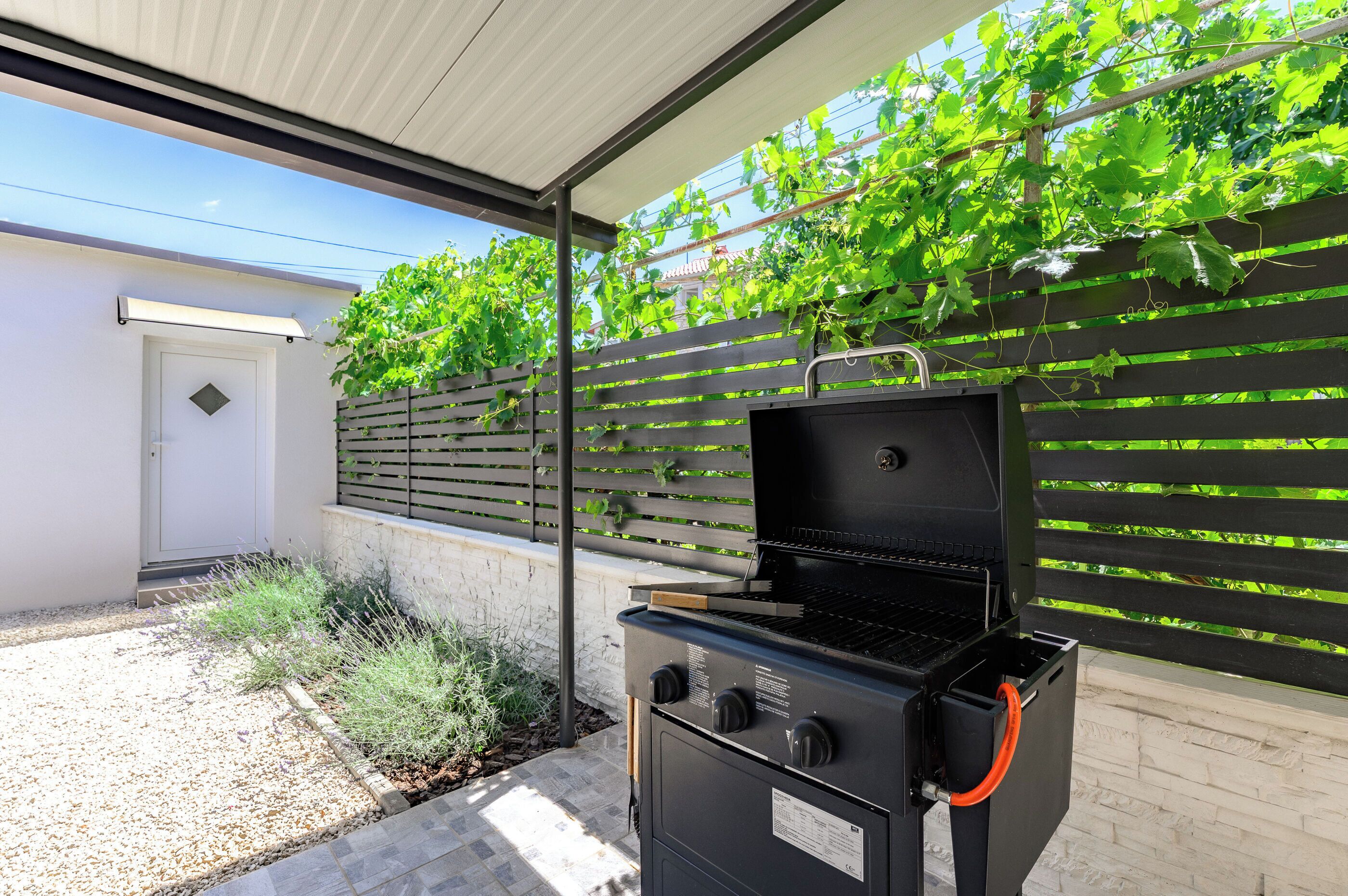An outdoor barbecue area with a grill, a wooden fence, and a white building in the background.