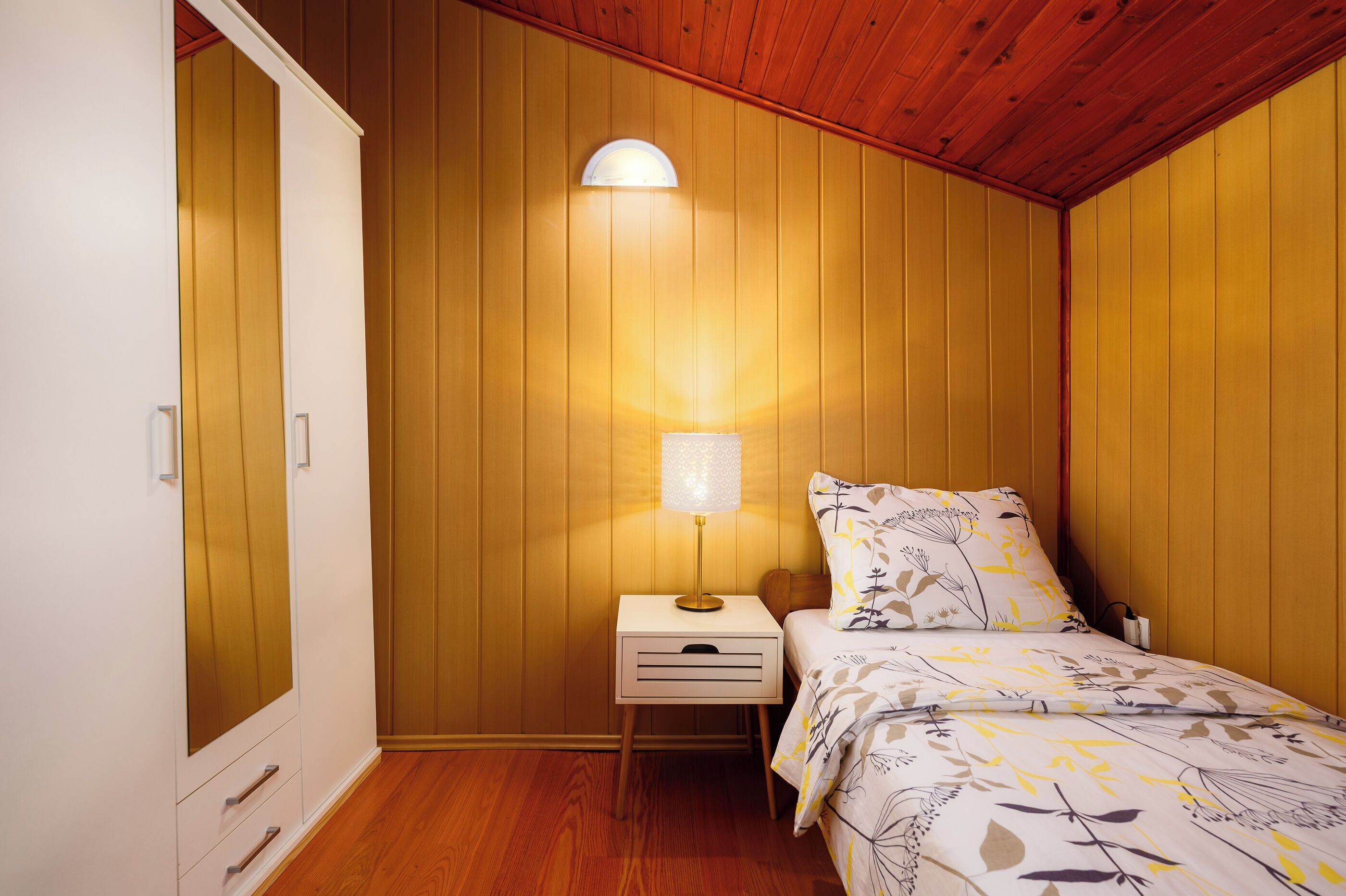 A bedroom with a wooden ceiling, a bedside lamp, a bed with floral bedding, and a wardrobe with drawers.
