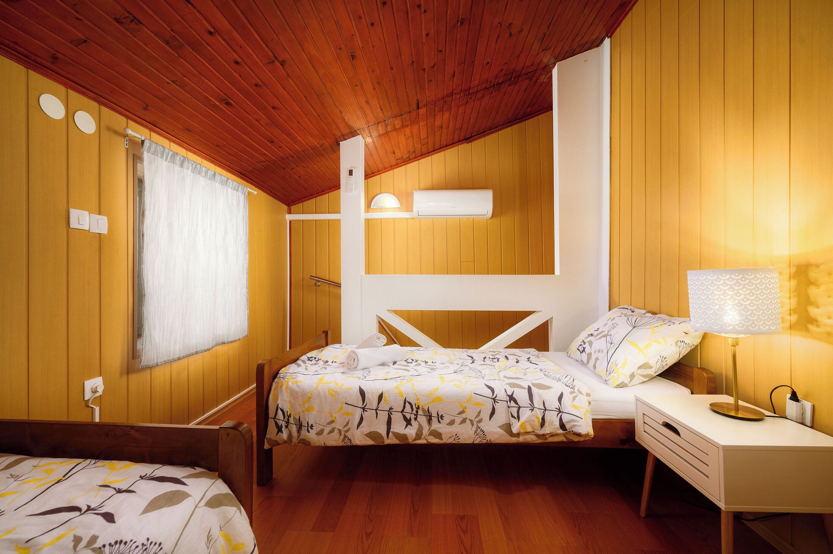 A cozy room with wooden walls and a sloped ceiling, featuring a bed with a floral bedspread, a bedside table with a lamp, and a window with curtains.