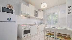 Eine kompakte Küche mit weißen Schränken, einem Einbauofen, einer Mikrowelle und einem Gasherd. Es gibt einen Esstisch mit Stühlen und ein Fenster mit durchsichtigen Vorhängen.