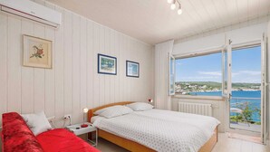 Ein Schlafzimmer mit einem Bett, einem roten Schlafsofa, einem Nachttisch und einem Fenster mit Meerblick.
