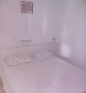 A simple, white room with a bed, two pillows, a small bedside table with a plant, and a framed picture on the wall. - House Garma (Blato)