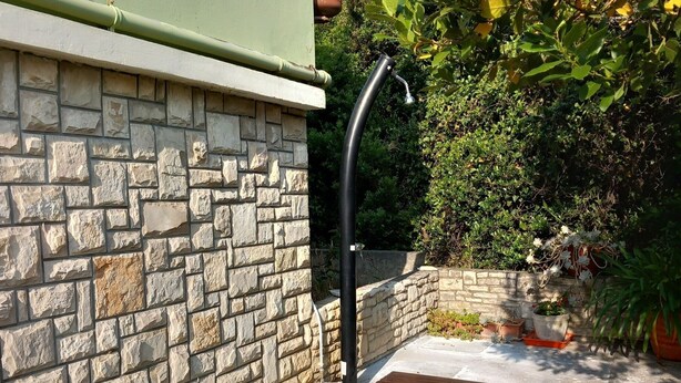 A stone wall with a green awning, a curved black pipe, and a small garden area with plants and a potted plant. - House Garma (Blato)