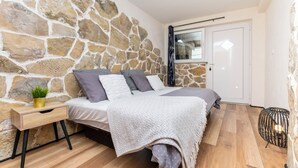 A bedroom with a stone wall, a bed with a white and gray blanket, a bedside table with a plant, and a small black lamp. - Apartment Golubinka (Dugi Rat)