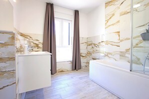 A bathroom with a marble wall, a bathtub, a sink, and a window with curtains. - Apartment Tin (Sveti Filip i Jakov)