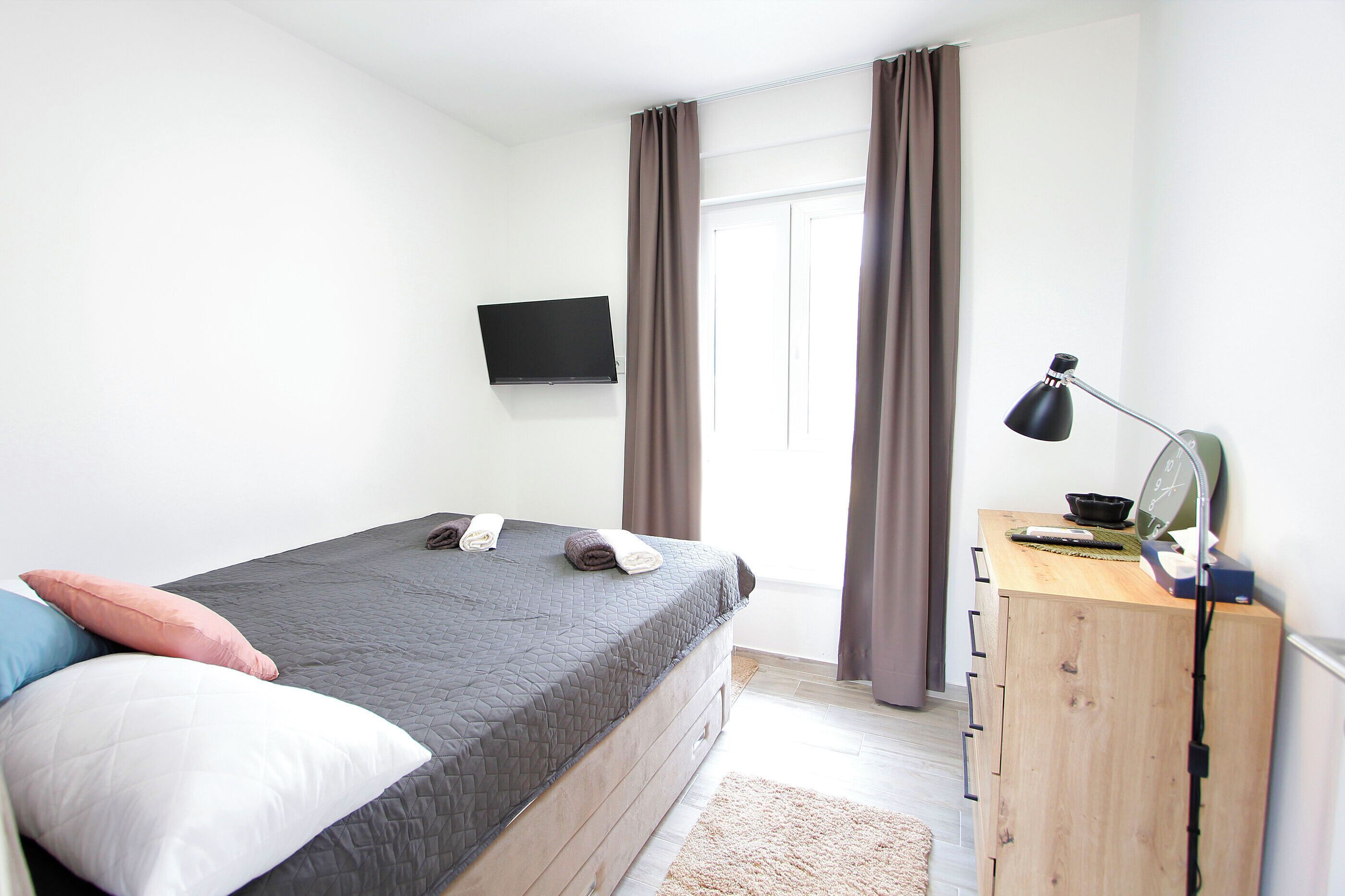 Quarto com cama, televisão na parede, escrivaninha com abajur e janela com cortinas.