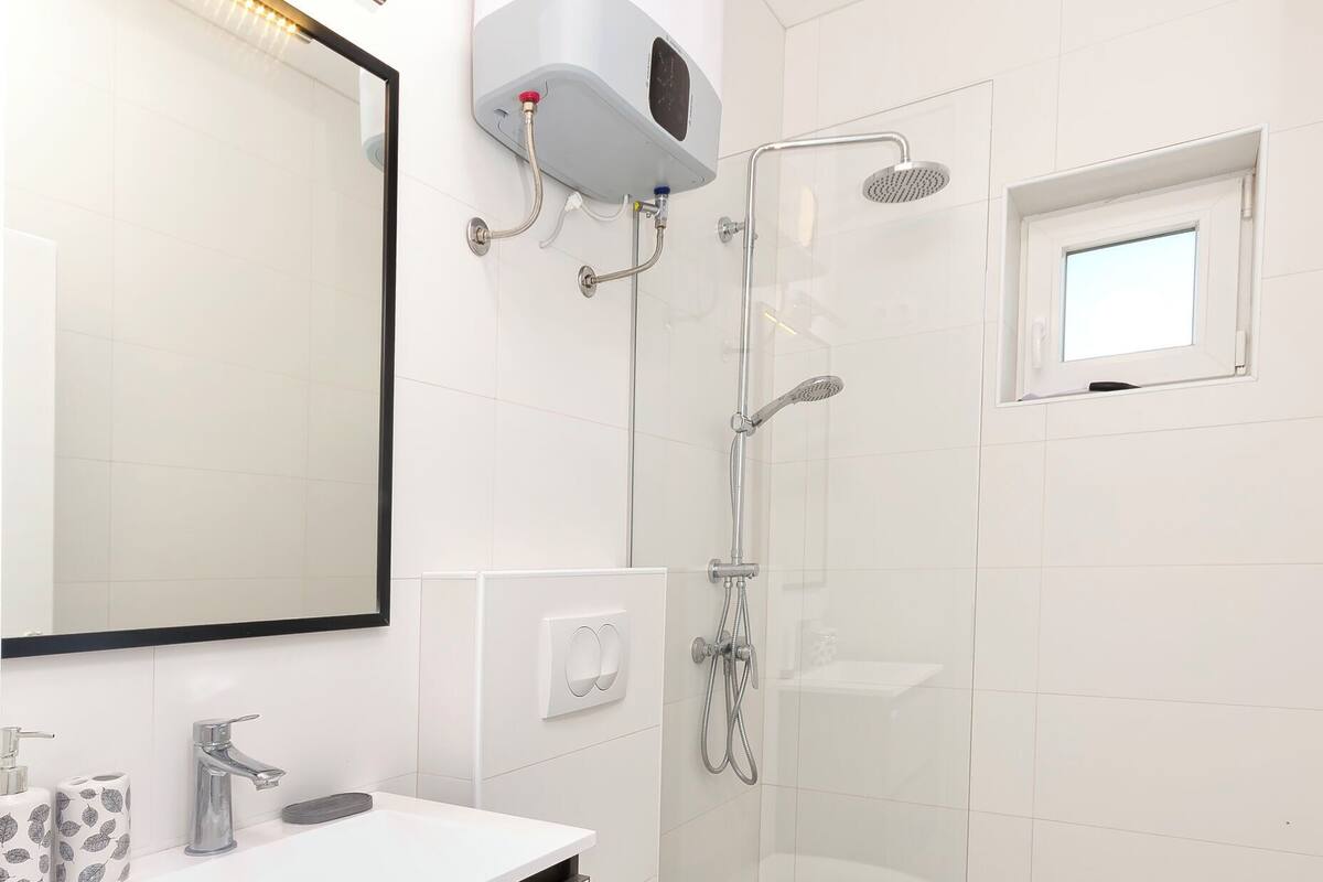 A modern bathroom with a white vanity, a toilet, a glass-enclosed shower, and a small window.