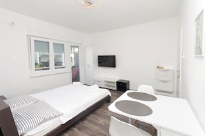 A modern, minimalist hotel room with a single bed, a small desk, a television, and a window with blinds. - Apartments Darinka (Vodice)