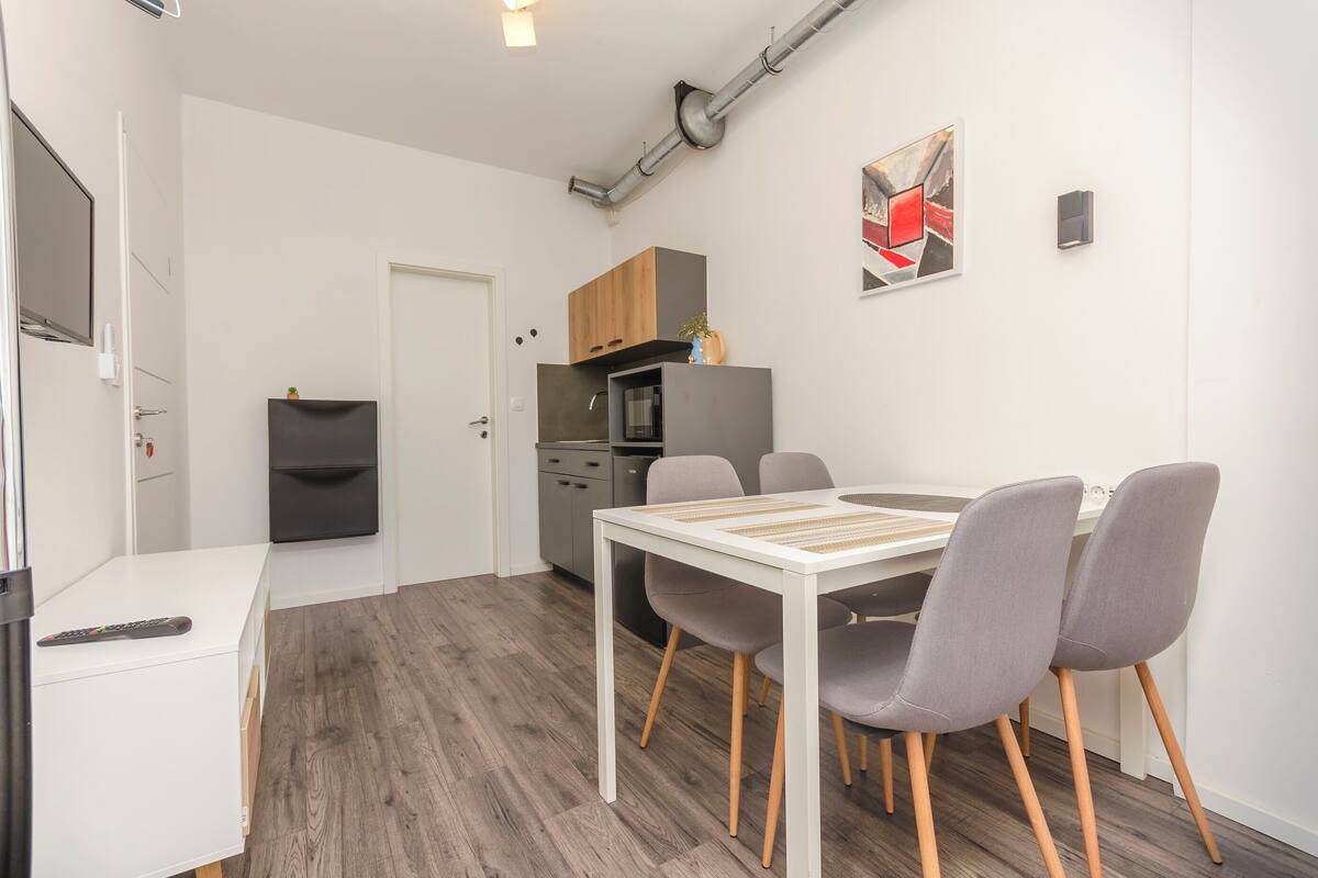 A compact kitchen with modern appliances, a dining area with a white table and grey chairs, and a small cabinet above the counter.