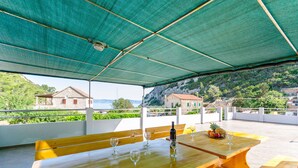 An outdoor terrace with wooden tables and benches, a green canopy, and a view of a hillside with buildings. - Apartment Kokic (Jelsa)