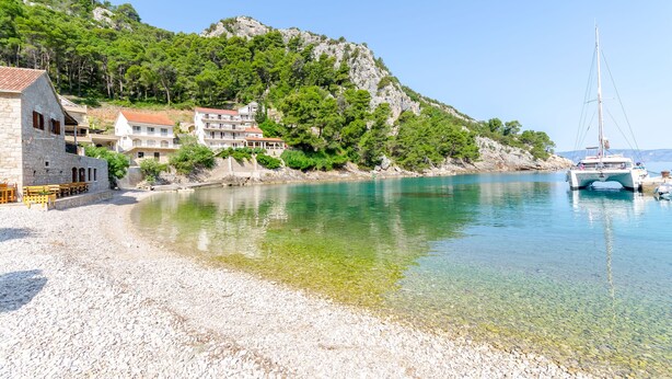 A coastal area with a pebble beach, clear water, a sailboat, and buildings on a hillside. - Apartment Kokic (Jelsa)
