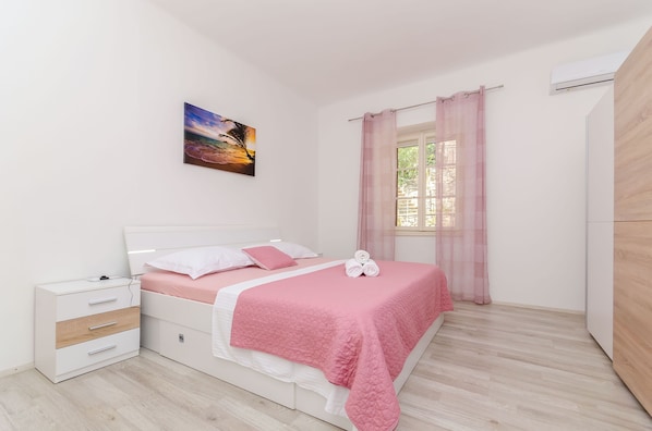 A bedroom with a bed, a nightstand, a dresser, a window with curtains, and an air conditioning unit. - Apartment Kokic (Jelsa)