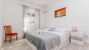 A bedroom with a bed, a chair, a bedside table, and a window with curtains. - Apartment Kokic (Jelsa)