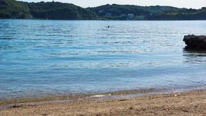 Uma praia com águas claras, pedras e uma colina distante com árvores e edifícios.