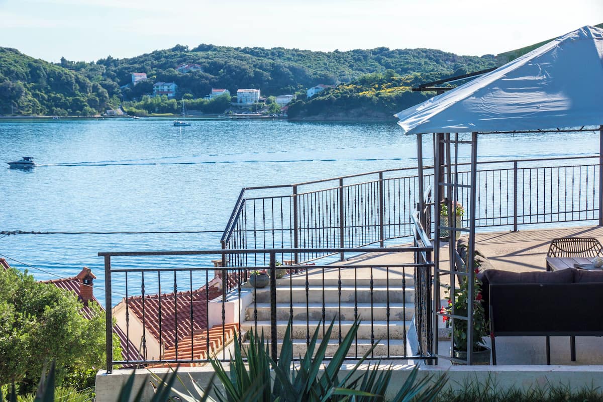A balcony with a view of a lake, a covered seating area, and steps leading down to the water.