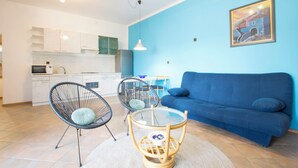 Uma sala de estar moderna com um sofá azul, duas cadeiras vazadas, uma mesa de centro de vidro e uma cozinha ao fundo.