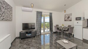 A modern living room with a flat-screen TV, a dining area with a table and chairs, a kitchen with a microwave, and a balcony with curtains. - Apartments Marija (Rogoznica)