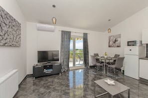 Ein modernes Wohnzimmer mit Flachbildfernseher, einer Essecke mit Tisch und Stühlen, einer Küche mit Mikrowelle und einem Balkon mit Vorhängen.