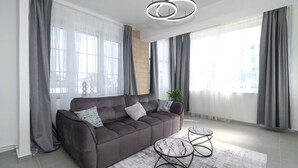 Uma sala de estar moderna com um sofá marrom escuro, duas mesinhas laterais e uma janela grande com cortinas leves.