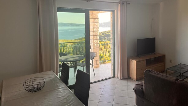 Ein Balkon mit Tisch und Stühlen, einem Fernseher auf einem Holzboden und Blick auf ein Gewässer und Grünflächen.