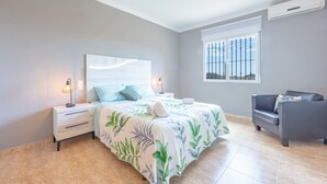A bedroom with a bed, nightstands, a chair, a window with bars, and an air conditioning unit. - Villa Pastor - Plusholidays (Calpe)