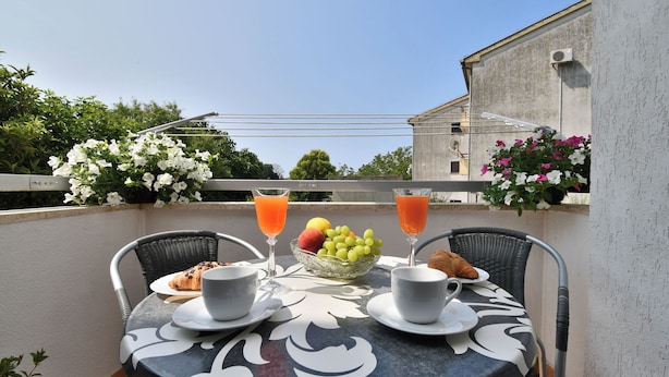 Een ronde tafel met een tafelkleed met bloemen, twee stoelen, een schaal met fruit, croissants en twee glazen jus d'orange.