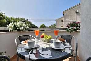 Een ronde tafel met een tafelkleed met bloemen, twee stoelen, een schaal met fruit, croissants en twee glazen jus d'orange.