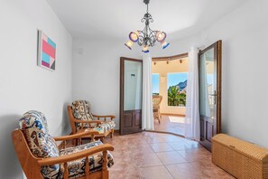 A room with two patterned chairs, a wicker bench, a chandelier, and a view of a balcony with outdoor furniture. - Villa Azur - Plusholidays (Calpe)