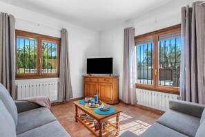 A room with a television, wooden cabinet, two sofas, a coffee table, and large windows with curtains. - Villa Azur - Plusholidays (Calpe)